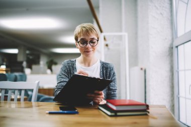 Pencerenin yanındaki masada oturan gülümseyen genç kadın, elinde not defterleri ve not panosuyla modern ofiste çalışırken kağıda rapor yazıyor.
