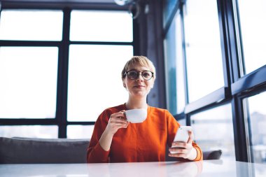 Düşük açılı, gözlüklü, kısa saçlı, gözlüklü, gülümseyen bir kadın kafeteryada bir fincan kahveyle oturuyor.