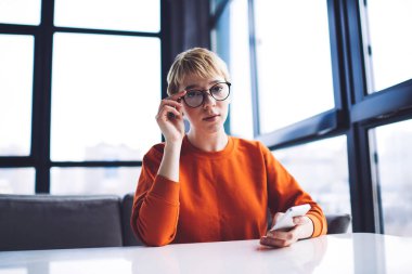 Düşük açılı, sıradan giyinmiş bir kadın pencerenin yanındaki masaya bakıyor. Mola sırasında cep telefonu kullanarak gözlüğe dokunuyor.