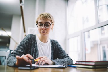 Ciddi kız öğrencilerin günlük kıyafetleri ve gözlükleriyle internette akıllı telefondan sörf yapması ve kütüphanede ödev yaparken not defterine notlar yazması.