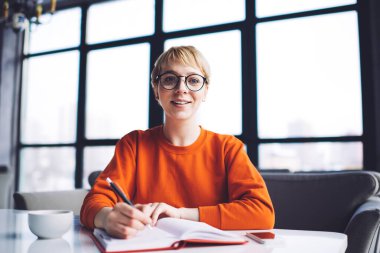 Genç serbest çalışan, dişlek gülümsemeli, elinde kahve fincanı ve yeni proje için fikirler yazan bir kadın.