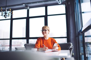 Turuncu kazaklı, gözlüklü, gülümseyen gözlüklü, akıllı telefon kullanan ve gündüzleri restoranda not defterine not alan olumlu bir kadın.