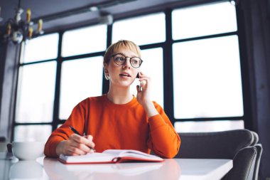 Düşük açılı genç bayan iş arayanın cep telefonuyla konuşması kahve fincanı ve defteriyle masada oturması.