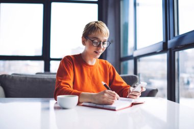 Günlük kıyafetlerle gülümseyen kadın serbest yazar pencerenin yanındaki masada oturmuş not defterine not alıyor ve uzaktan çalışırken akıllı telefona göz atıyor.