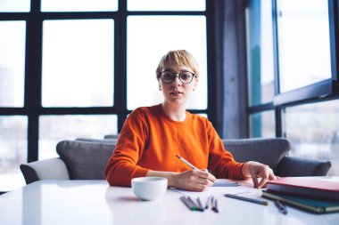 Ciddi kadın serbest çalışan, gündelik kıyafetlerle pencerenin yanındaki masaya bakan, kahve fincanı taşıyan, proje üzerinde çalışırken not alan biri.
