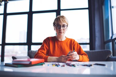 Kısa saçlı, gündelik kıyafetli, masa başında renkli kalemlerle oturan genç ve düşünceli bir kadın işçiden.