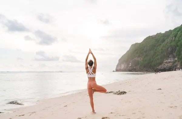 Back view full length of anonymous barefooted fit female in sportswear performing Tree with Arms Up yoga asana on spectacular sandy beach