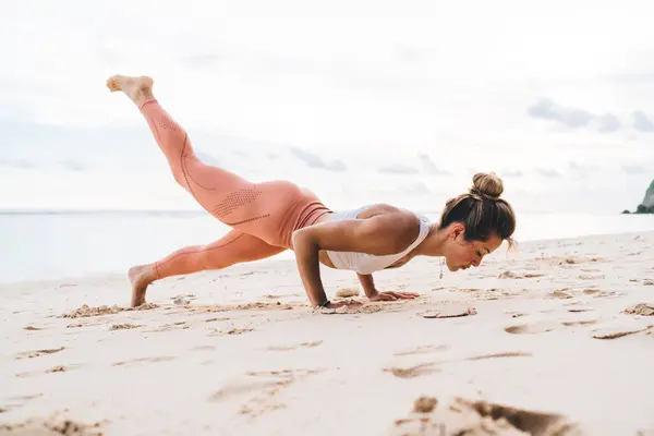 Side view of flexible female in activewear practicing Four Limbed Staff pose and lifting leg up on sandy beach near calm ocean in daytime on