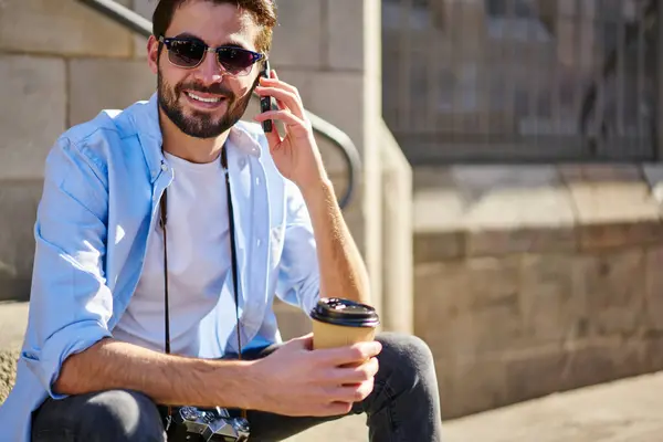 カジュアルなウェアで陽気な男性の観光客は 庭で休んでコーヒーを飲んでいる間 明るい笑顔で携帯電話で話すカジュアルなカメラで ストックフォト