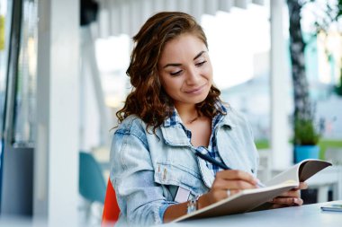 Gündelik giyinen yetenekli bayan öğrenci sokak kafeteryasında dinleniyor ders kitabında eğitim bilgisi yazıyor, yaratıcı Kafkasyalı hippi kız en sevdiği hobi için not defterinde skeçler çiziyor.