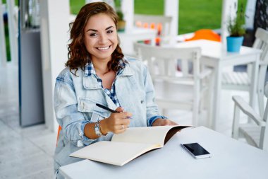 Sokak kafesinde planlama sırasında kameraya gülümseyen günlük kıyafetli neşeli kız öğrencinin portresi, ders kitabı ve akıllı telefon cihazlı komik milenyum blogcusu yayınlama metni yaratıyor