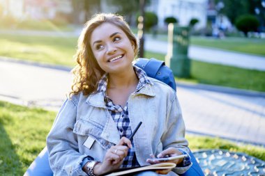 Modern cep telefonu ve ders kitabı olan neşeli bayan öğrenci, güneşli bir günde gülümsemek için ders kitabı, akıllı telefonlu çekici blogcu parkta iyi vakit geçirmenin keyfini çıkarıyor.