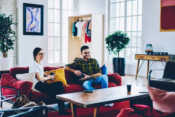 Cheerful Caucasian hipster girl and happy Middle Eastern man enjoying live communication during coffee break in comfy living room, funny male and female friends with takeaway cups smiling indoors
