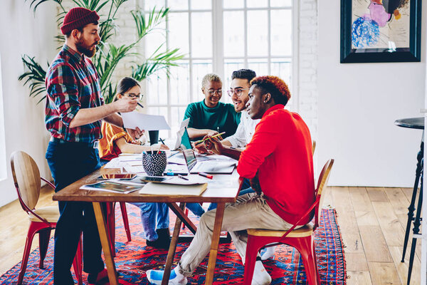 Caucasian man with paper report talking about business strategy while cooperating with diverse colleagues, professional hipster guy communicate with multiracial partners about graphic design