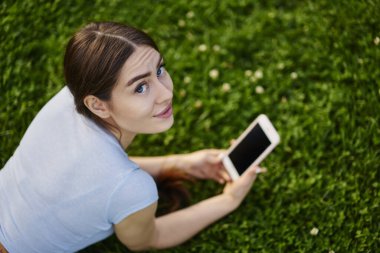 Elinde dijital akıllı telefon tutan ve boş zamanlarında kameraya bakan beyaz bir kadının portresi modern cep telefonu cihazıyla parktaki yeşil çimlerde poz veren bir yeniyetme kız.