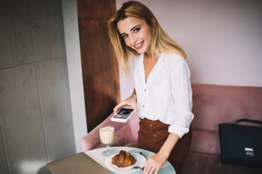 Zarif kıyafetli bir kadın cep telefonuyla kruvasanın fotoğrafını çekiyor. Bir bardak içki ve netbook masada, kanepede dikilip kameraya bakıyor.