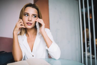Beyaz bluzlu, düşünceli genç bir bayan masada oturuyor ve not defterine not alıyor evde akıllı telefonuyla konuşurken başka tarafa bakıyor.