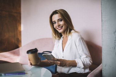 Resmi giysiler içinde genç ve çekici sarışın bir kadın. Masada oturmuş kahve ısmarlıyor ve gülümsüyor. Modern ışık ofisinde akıllı telefonları inceliyor.