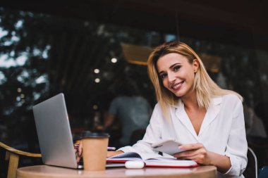 Cep telefonuyla gülümseyen ve netbook ile çalışan neşeli genç bir bayan sokak kafesindeki masada kameraya bakıyor.