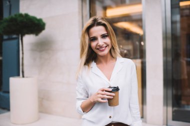 Beyaz bluzlu, karizmatik bir kadın gülümseyen ve kameraya bakan modern binanın girişindeki şehir caddesinde kahve keyfi yapan.