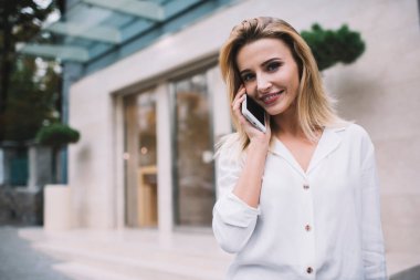 Beyaz bluzlu kadın akıllı telefonla konuşuyor ve modern binanın girişinde dikilirken kameraya bakıyor.