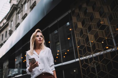 Beyaz bluzlu, düşünceli, genç, sarışın bir kadın cep telefonundan mesaj okuyor. Modern binanın yanında duruyor.