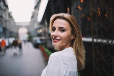 Pozitif gülümseyen genç sarışın bir kadının akşamları caddede modern binanın yanında dikilip arkasını kollamasının yan görüntüsü.