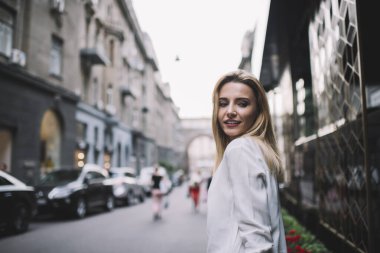 Beyaz gömlekli genç bayan şehirde kaldırımda yürüyor. Gündüz vakti binaların ve arabaların cephelerinin yanında ve başka tarafa bakıyor.