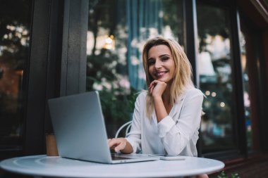 Neşeli kadın serbest çalışan kameraya bakıyor ve masaya yaslanıyor ve online çalışma sırasında netbook 'ta yazıyor.