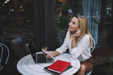 Masada dizüstü bilgisayarla otururken gözleri kapalı müzik dinleyen ve cep telefonunu karıştıran pozitif kadının yan görüntüsü