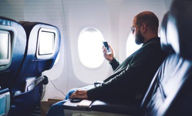 Ciddi yetişkin bir adamın gözlük takıp, günlük giysiler giyerken akıllı telefon kullanması ve gündüz vakti yolcu koltuğunda oturması.