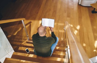 Yukarıdan bakan adam boş sayfalarla ahşap kitap okuyor. Gündüz vakti modern bir dairede vakit geçiriyor.