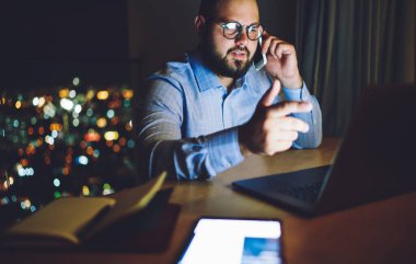Düşünceli erkek girişimci geceleri çalışırken dizüstü bilgisayarın ekranını işaret ederken cep telefonuyla konuşuyor.