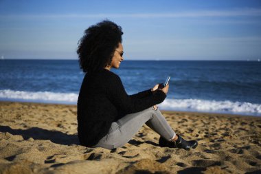 Sahilde oturup akıllı telefon sörfü yapan gündelik giysili ve güneş gözlüklü Afro-Amerikan kadın.