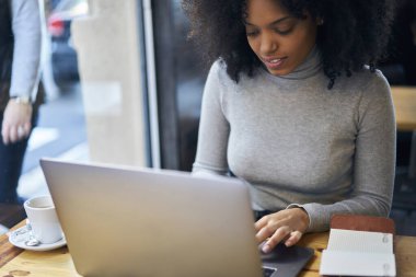 Yoğunlaşmış, günlük giysiler içinde genç bir kadın ahşap masada oturmuş bilgisayarın klavyesinde uzaktan çalışırken daktilo yazıyor ve bir fincan kahve içiyor.