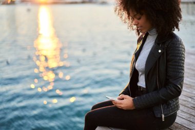 Şık saçlı, odaklanmış genç Afrikalı Amerikalı turist gün batımında göl kenarında rıhtımda uzanırken akıllı telefon takıyor.