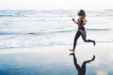 Aktif kıyafetli genç bir kadın sabah kardiyo antrenmanında koşu hedeflerine ulaşır. Zayıf, koyu tenli, enerjik, açık denizde koşan atletik gücü eğiten bir kadın.