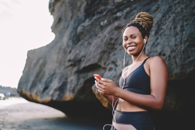 Eşofmanlı neşeli bayan koşucu, sabah dışarıda antrenman yaparken kendini iyi hissediyor. Elektronik kulaklıkla motivasyon şarkıları dinleyen mutlu fit bir kızın yarım boy portresi.