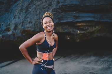 Cep telefonunda müzik uygulamasından gelen ses şarkılarını dinlerken gülümseyen mutlu Afro-Amerikan kadın portresi. Neşeli koyu tenli koşucu sportif bluz giymiş, sabah keyfine bakıyor.