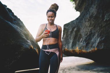 Mobil teknolojili çekici bir kız. Sahil şeridinde sabah antrenmanı yaparken medya içeriğini kontrol ediyor. Elektronik kulaklıklı mutlu bir bayan sporcu sosyal ağlarda akıllı telefon mesajlarıyla eğleniyor.