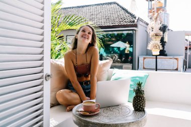 Bali 'de dizüstü bilgisayarla rahat bir koltukta otururken rahat bir şekilde gülümseyen, gülümseyen bir kadın.