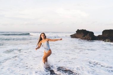 Güzel bir kadın, beyaz mayo giyer, deniz kıyısında yürür, açık havada köpüklü deniz dalgalarıyla yıkanır ve kameraya bakar.