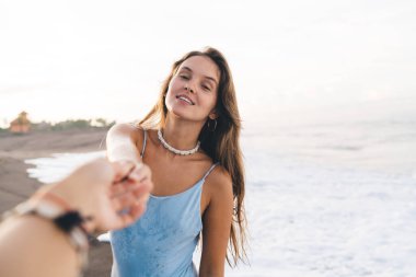 Mavi mayo giyen genç ve güzel bir bayan kumlu ve ıslak deniz kıyısında kameraya bakarken erkek arkadaşlarını nazikçe tutuyor.