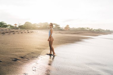 Sıska, çıplak ayaklı, şık bir mayo giymiş bir kadının yaz tatilinde kumlu sahilde tatil köyünde duruşu.