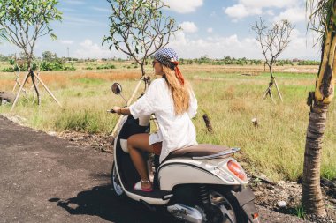 Kırsal kesimde kırsal kesimde beyaz scooter kullanan, uzun saçlı, uzun saçlı, tanınmayan genç bir kadın gezginin yan görüntüsü.