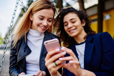 Gündüz vakti arka planda binayla birlikte sokakta bankta otururken günlük kıyafetlerini giymiş, telefonları kontrol eden pozitif, çok ırklı genç bayan arkadaşlar.