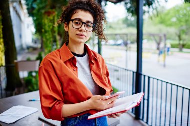 Arzu dolu genç bir kız öğrenci kameraya bakarken planlayıcıyla duruyor ve sokak kafeteryasında ödev yapmak için akıllı telefonu kullanıyor.