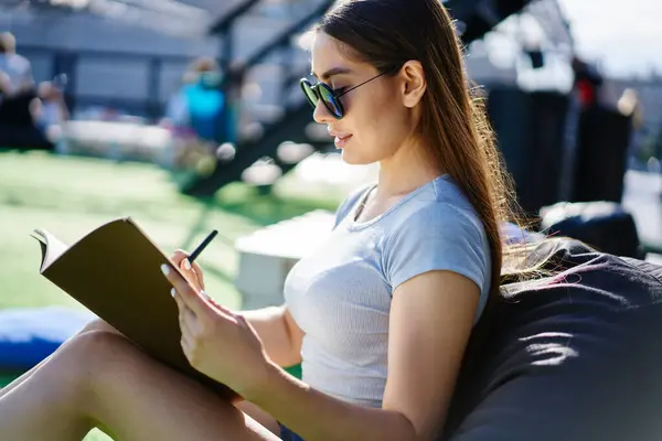 Talented female designer with brunette hair creating sketches during sunny day in campus,millennial hipster girl in trendy sunglasses learning and studying outdoors sitting in bag chair and doing test