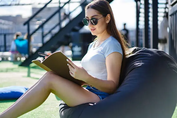 Talented female designer with brunette hair creating sketches during sunny day in campus,millennial hipster girl in trendy sunglasses learning and studying outdoors sitting in bag chair and doing test