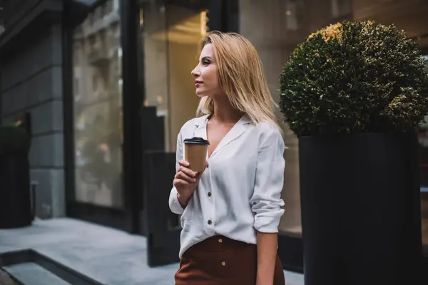 Unemotional young female in white blouse with rolled up sleeves drinking coffee in disposable cup while standing near shop entrance and looking away